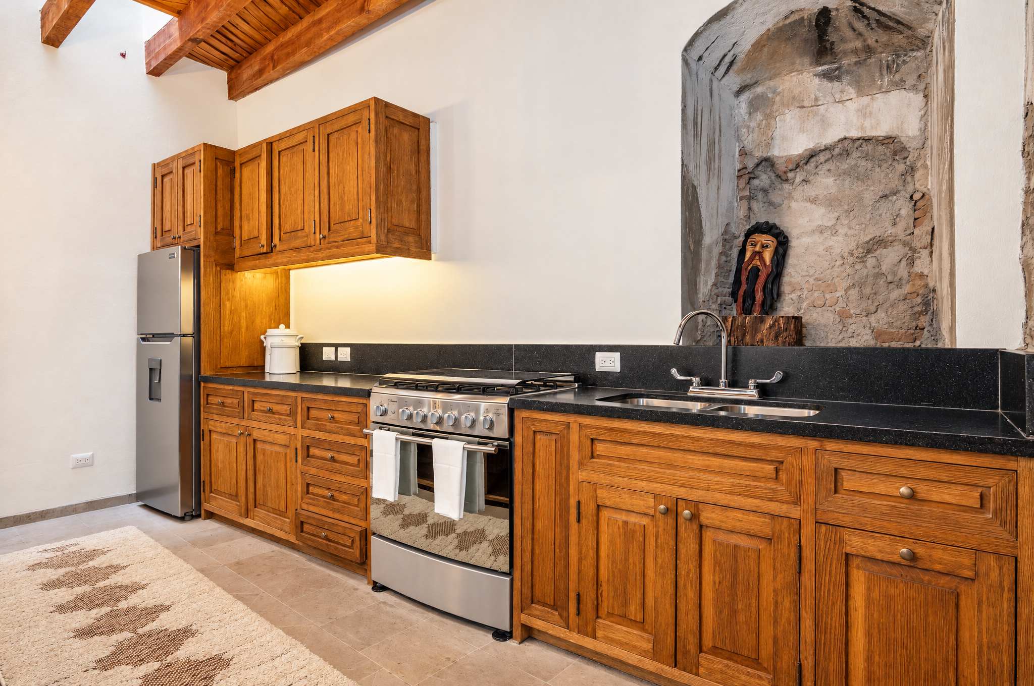 Cocina con gabinetes de madera, encimeras negras, estufa de acero inoxidable y vigas de madera a la vista