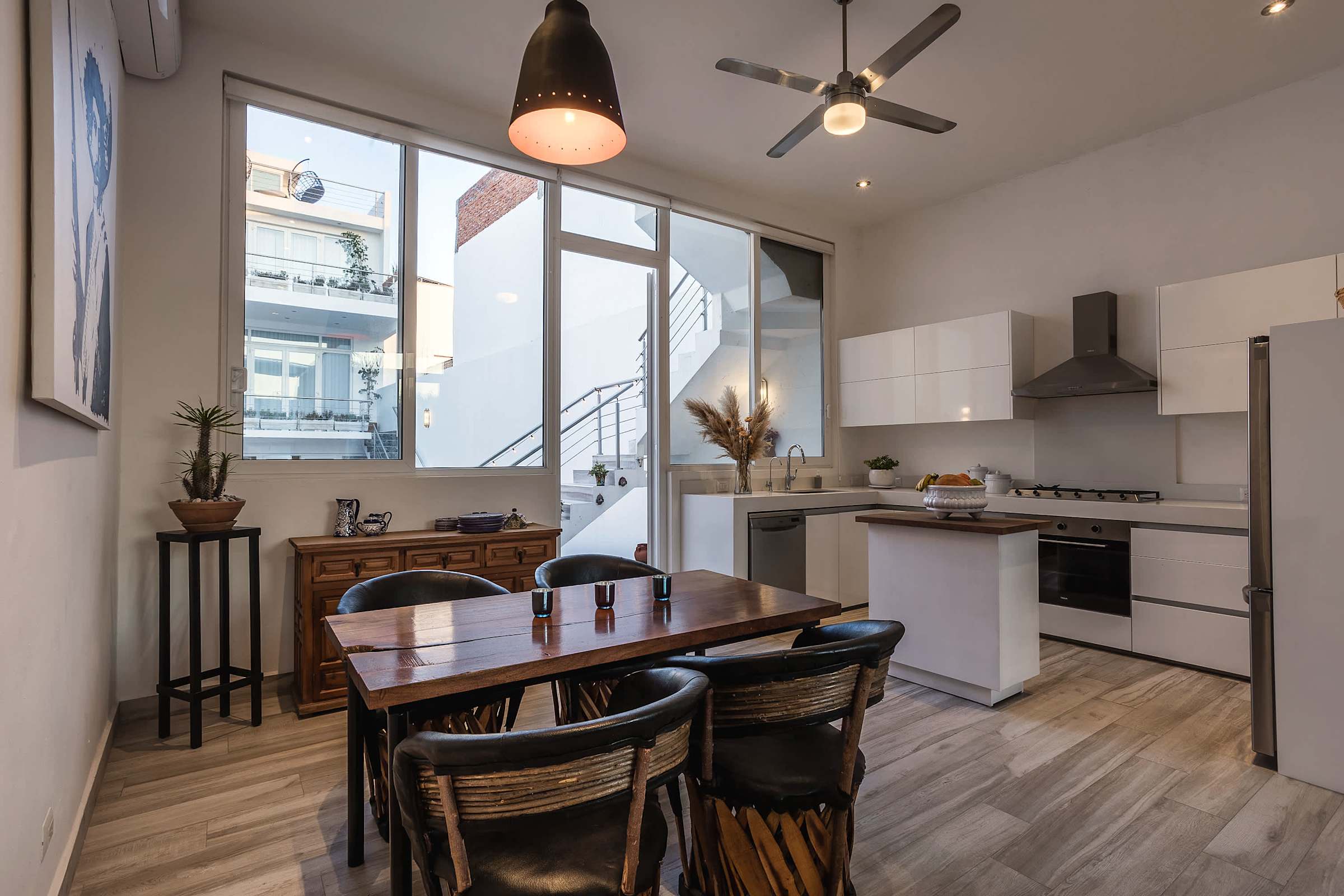 Cocina y comedor con mesa de madera, gabinetes blancos y grandes ventanales con vista a la azotea
