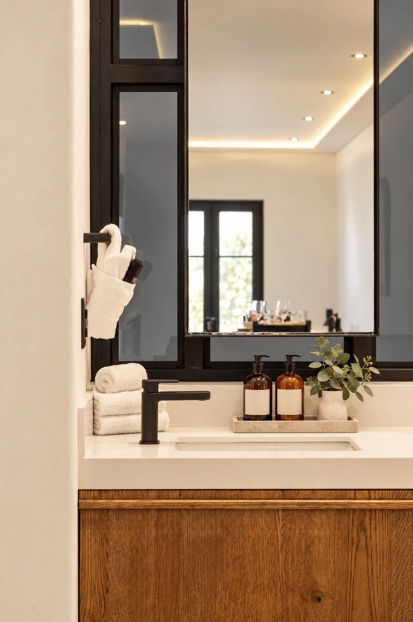 Baño con espejo grande, grifería negra, frascos de vidrio ámbar y iluminación cálida en el techo