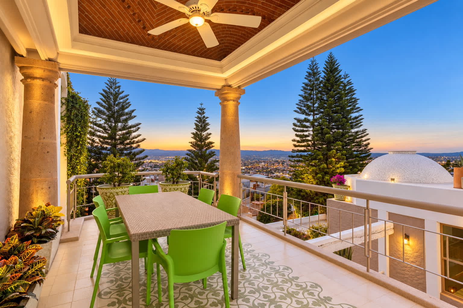 Covered terrace with dining table, green chairs, and panoramic city views at sunset