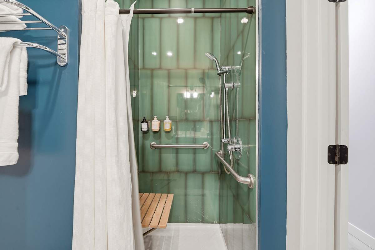 Step into the accessible walk-in shower showcasing sage green vertical subway tile, a wooden bench seat, stainless steel grab bars, and a hand-held shower head.