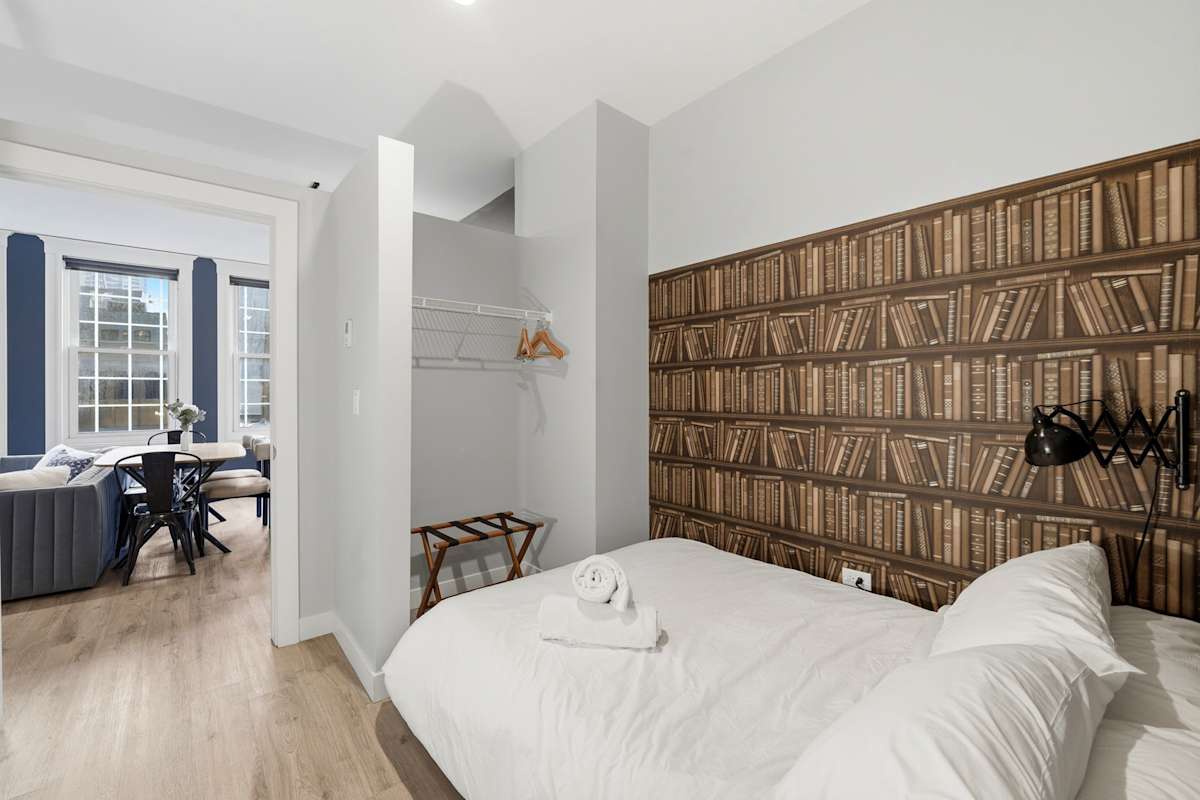 View the bookshelf-themed bedroom looking out toward the main living area, featuring an open closet niche with luggage rack and wall-mounted reading lamp.