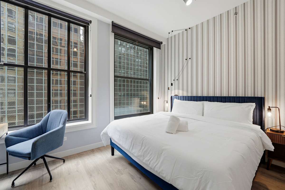 Rest in the bedroom styled with a blue upholstered king bed frame, vertical striped feature wallpaper, blue office armchair, and large city-facing windows.