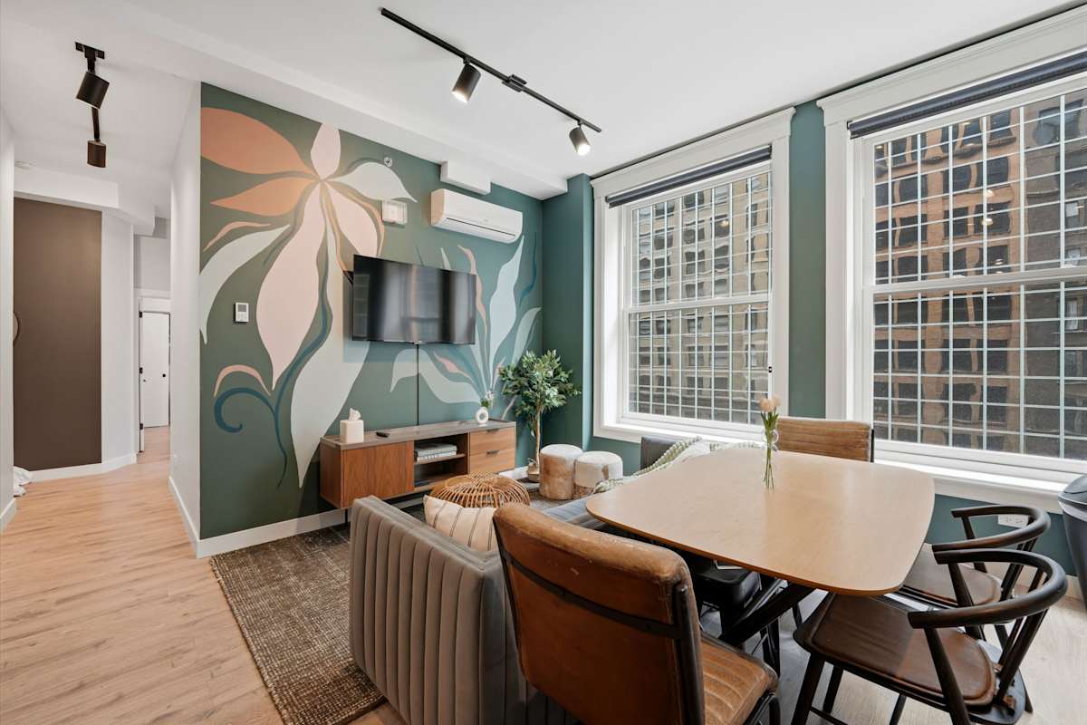View the modern open-plan living and dining space featuring a large floral wall mural, channel-tufted sofa, wooden dining set, and large grid windows.