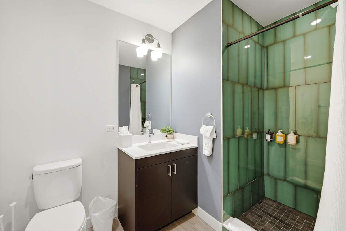 View the ensuite bathroom setup showing the dark vanity unit, chrome fixtures, toilet, and walk-in shower lined with sage green vertical subway tiling.