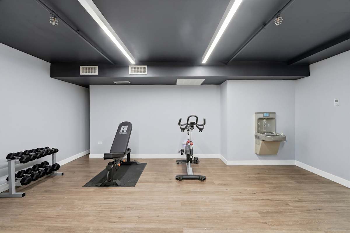 Train in the secondary fitness area featuring light wood-look flooring, a dumbbell rack with hand weights, an adjustable weight bench, a stationary exercise bike, and a wall-mounted water fountain.