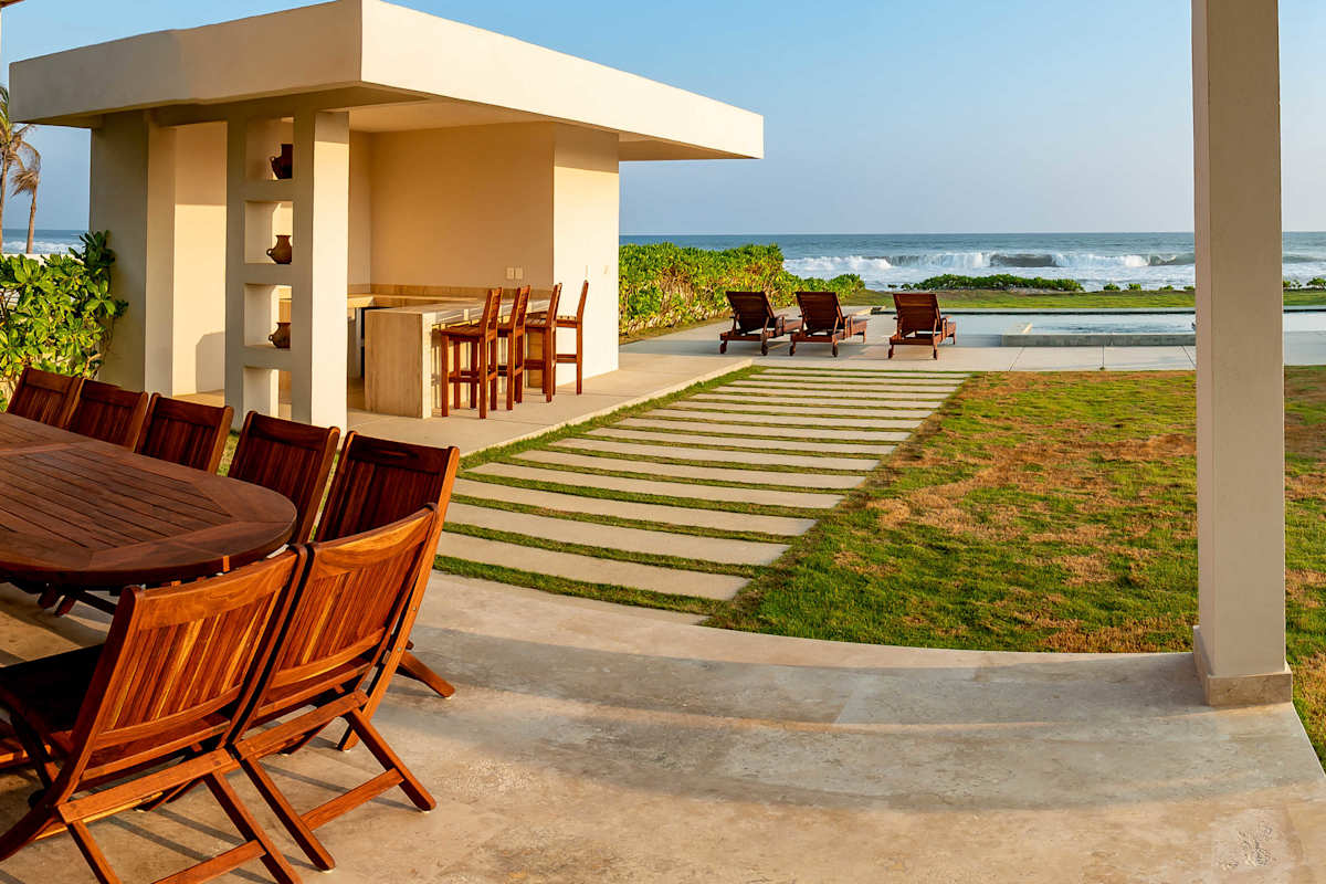Outdoor wooden dining set on a spacious garden terrace with a manicured lawn and pool area in the backdrop
