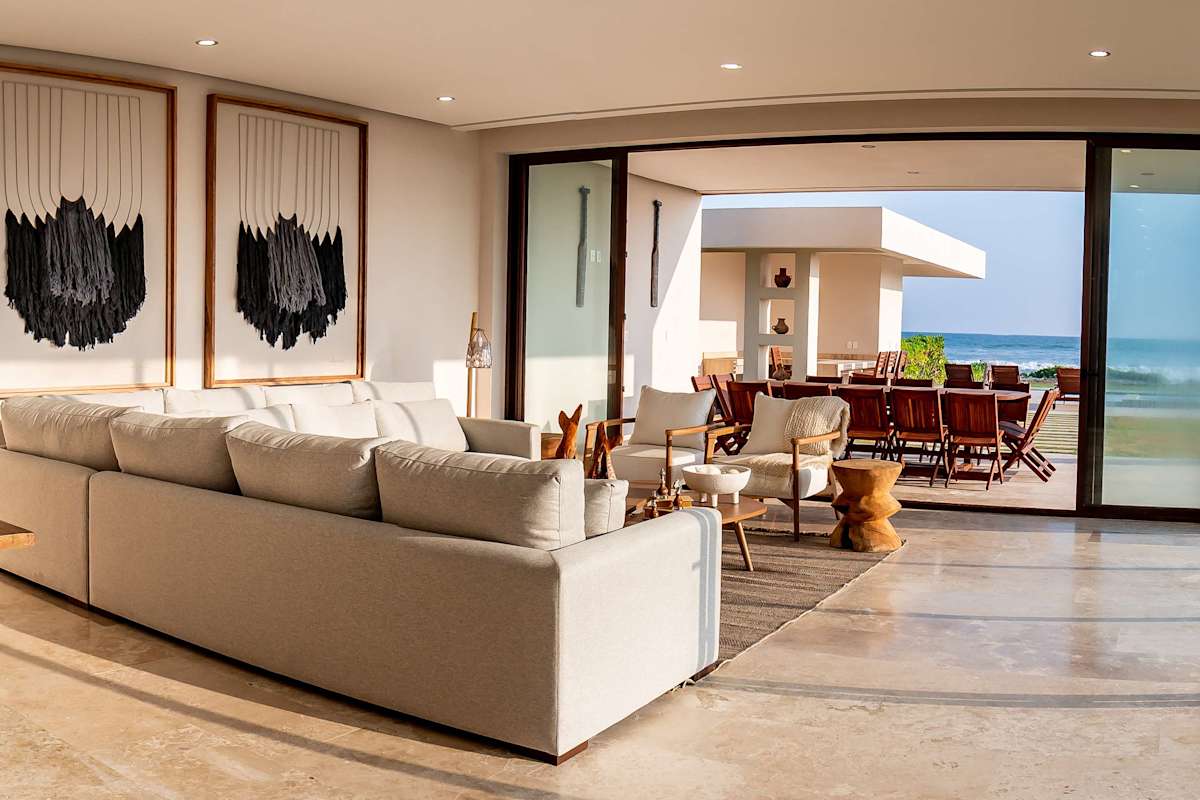 Open-plan living area with floor-to-ceiling glass doors, modern sofas, and direct ocean views — bring the outdoors in