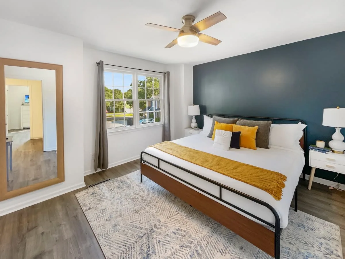 A hotel room with a large bed, ceiling fan, window, mirror, and rug.