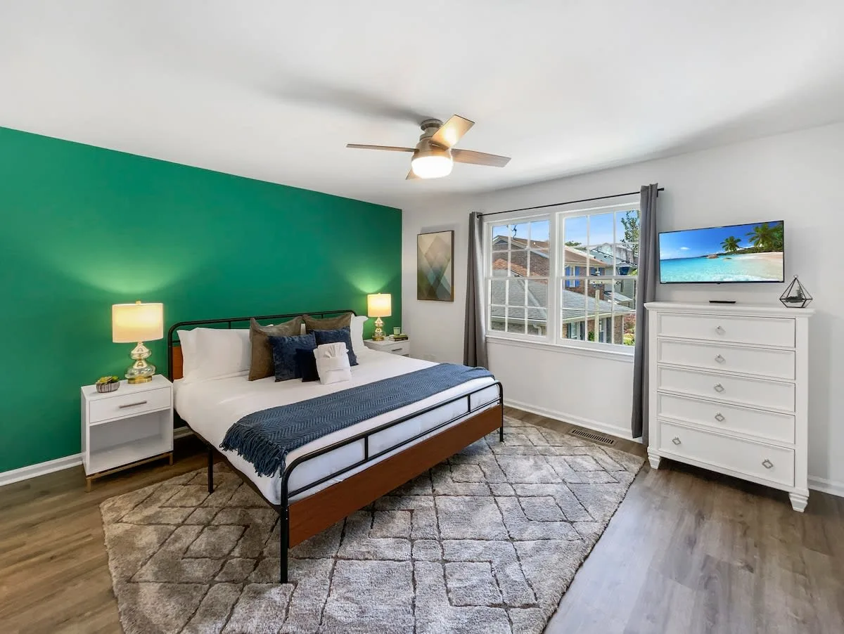 A wide view of a bedroom with a green accent wall, a bed, a TV on a dresser, and a window.