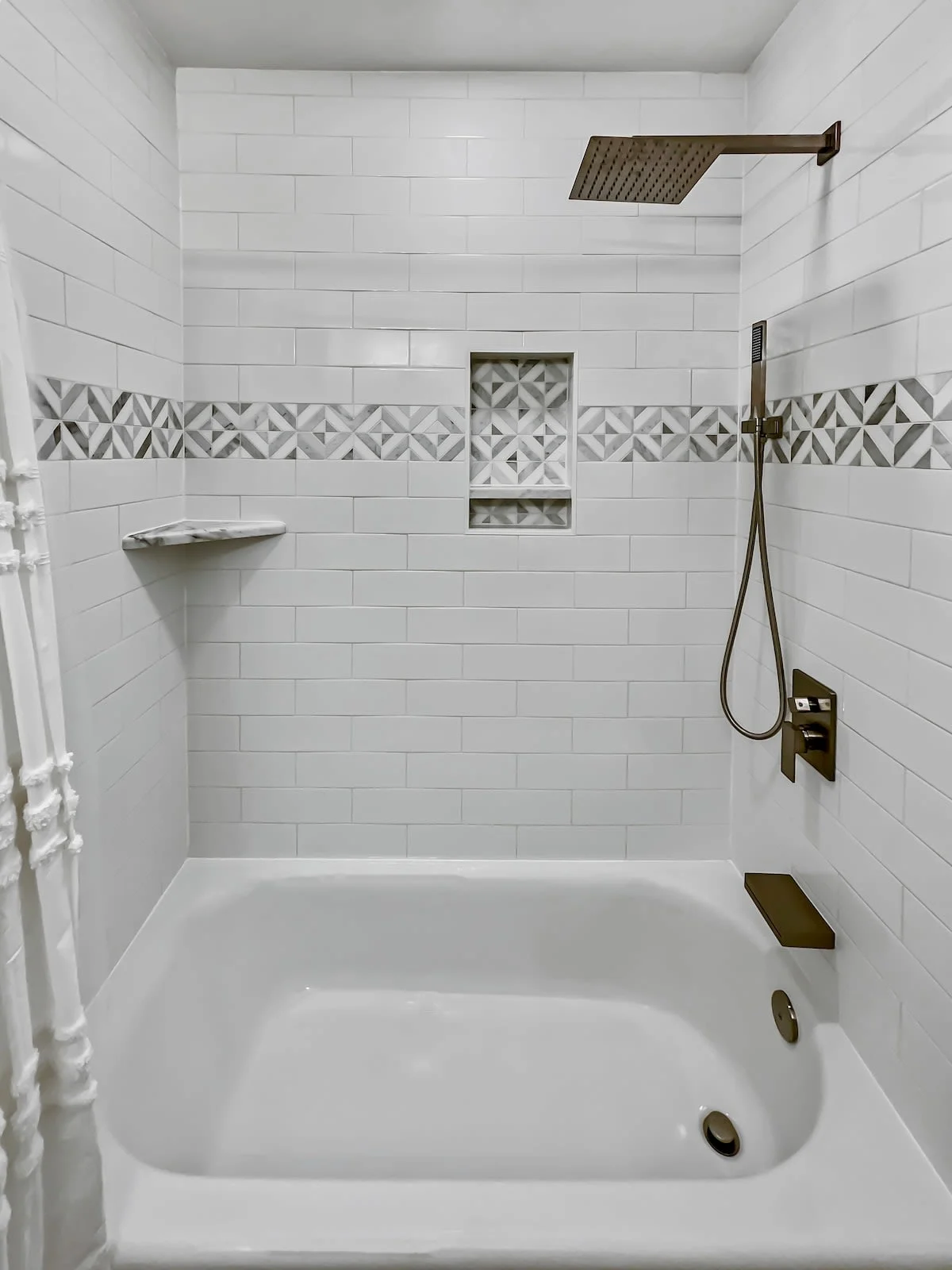A clean and modern bathroom with a white bathtub and shower, tile walls, and bronze fixtures.
