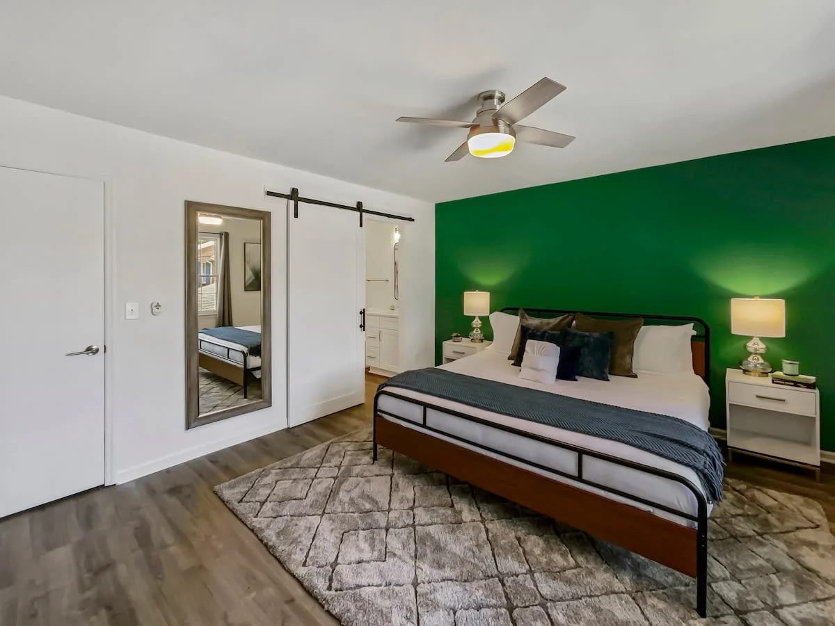 A wide-angle view of a bedroom with a green accent wall, a bed with white and dark-colored bedding, two nightstands with lamps, a large mirror, and a ceiling fan.