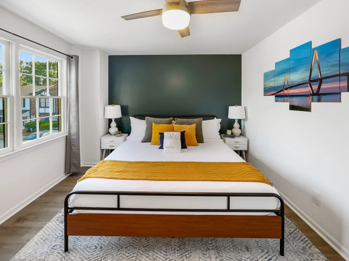A neatly made bed with yellow and white pillows and a yellow blanket in a room with a window, two nightstands, and artwork on the wall.