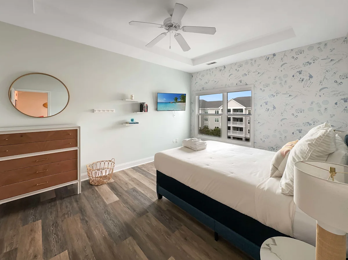 A clean and bright room with a bed, dresser, mirror, ceiling fan, window, and TV.