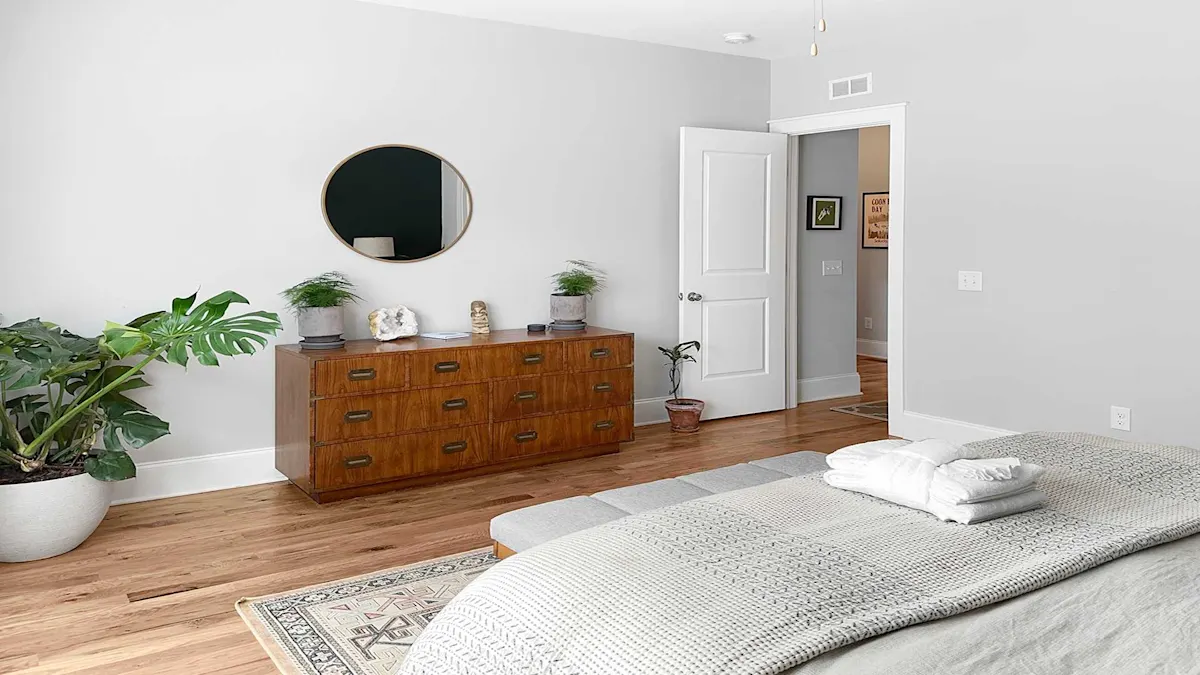 A neatly arranged bedroom with a wooden dresser, round mirror, and potted plants, featuring a bed with folded linens on a hardwood floor.