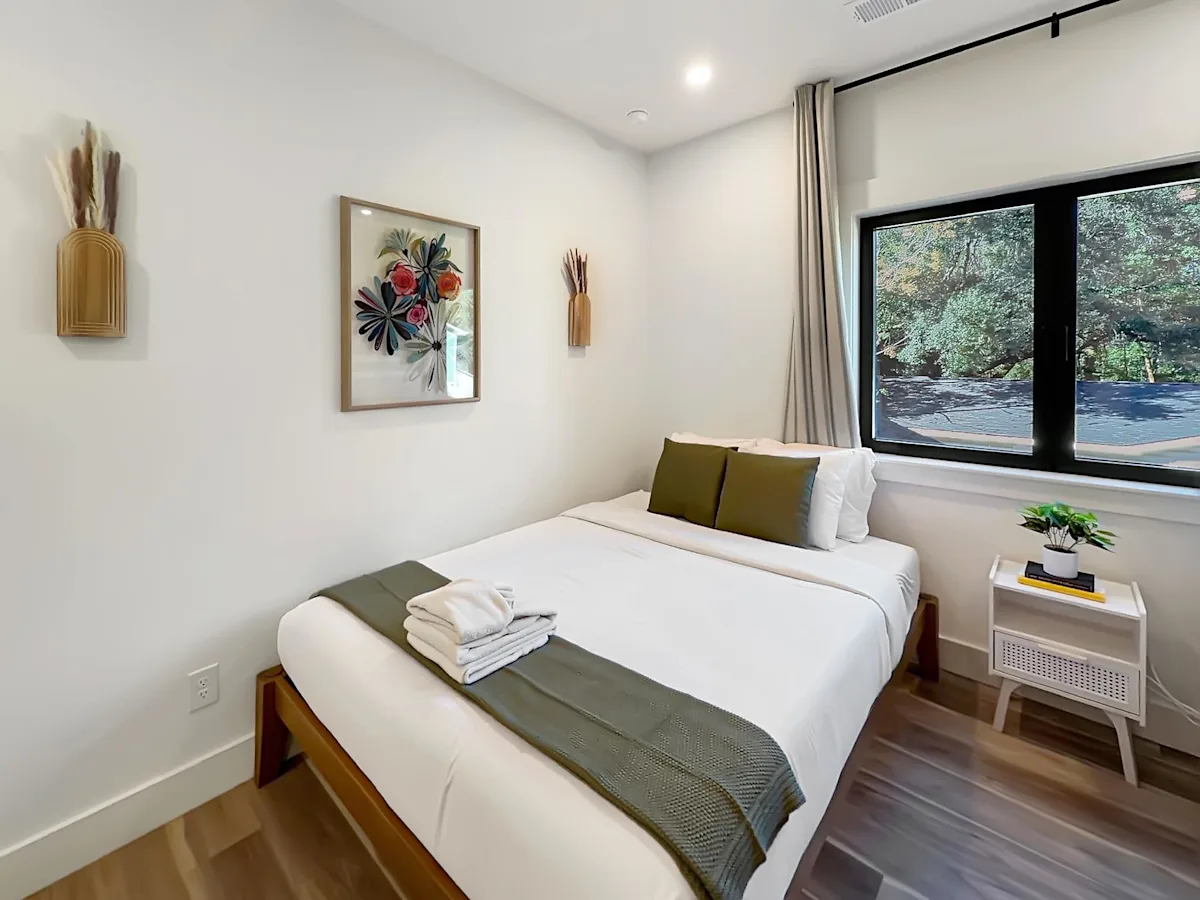A neatly made bed with white sheets and green accents in a room with white walls and a large window.