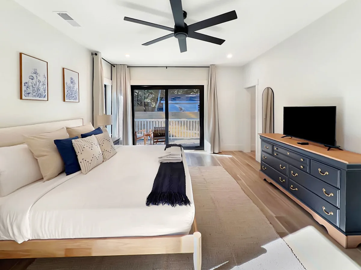 A large bedroom with a king-sized bed, a TV on a dresser, and a sliding glass door leading to a balcony.