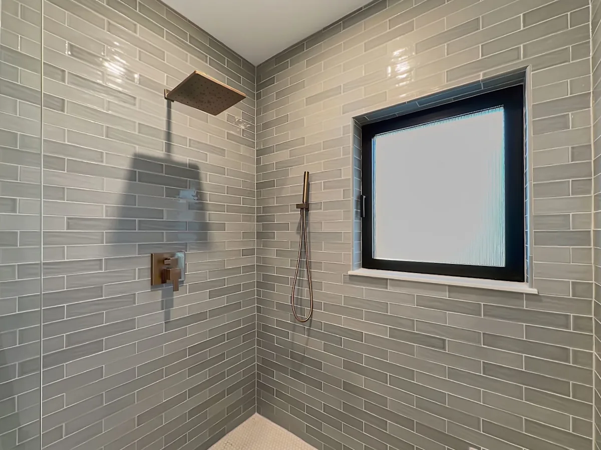 A shower with gray tile walls, a rainfall shower head, and a window with a black frame