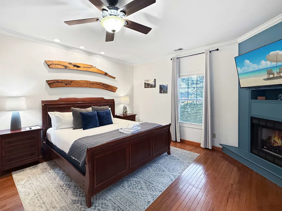 A wide view of a bedroom with a dark wood bed frame, nightstands, and a TV mounted above a fireplace.