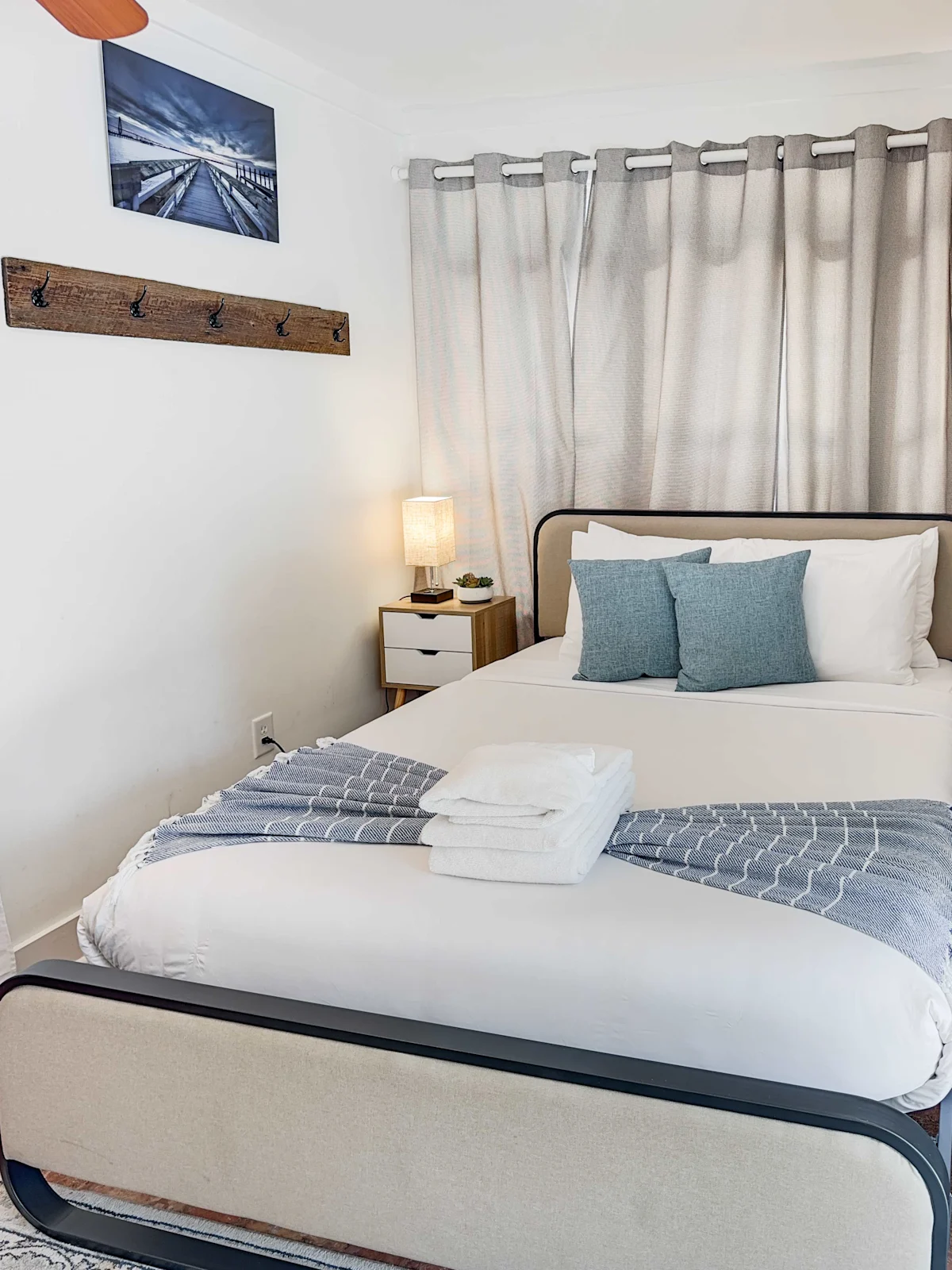 A neatly made bed with white and blue linens, a nightstand with a lamp, and a window with curtains.
