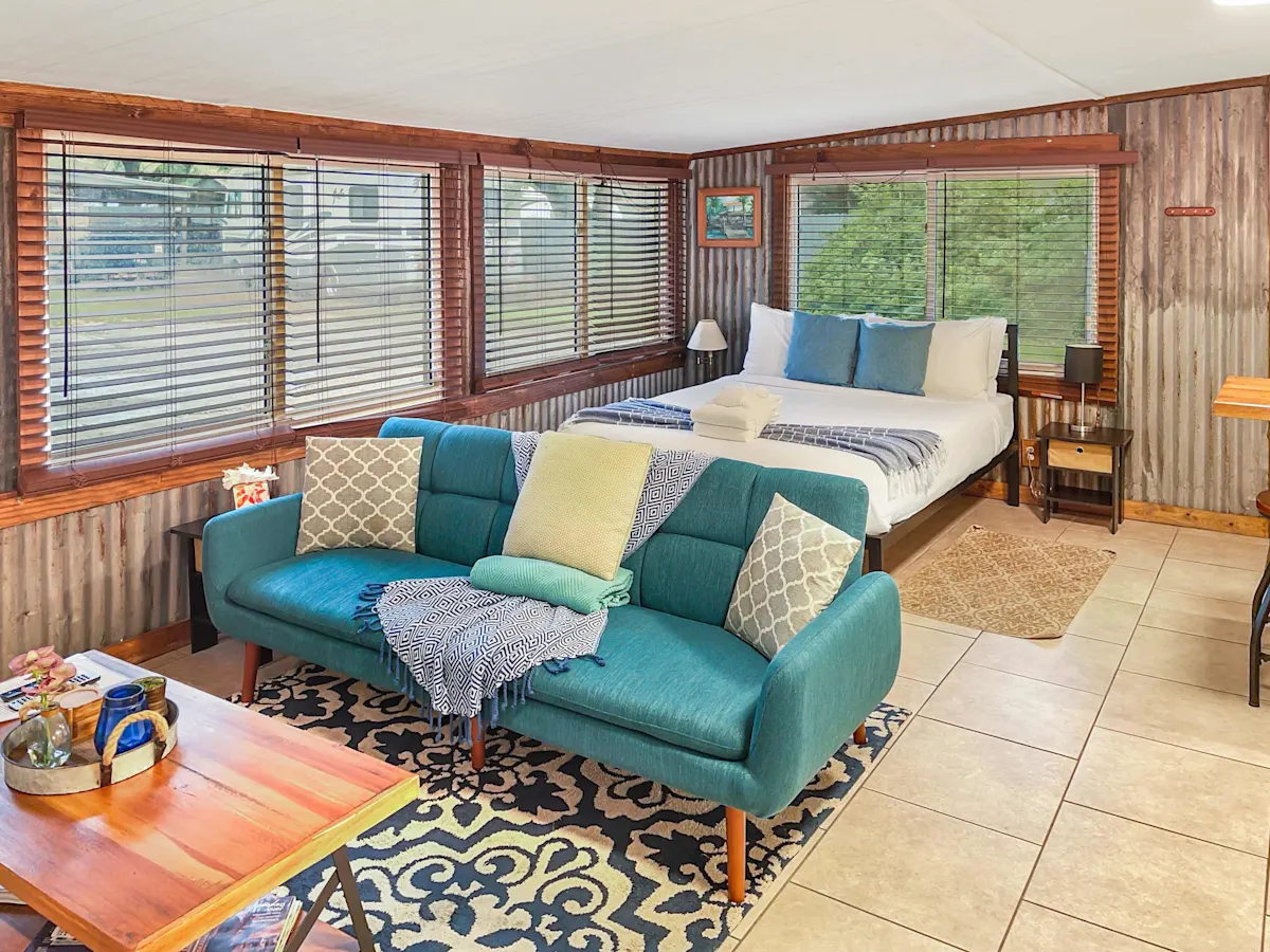 A cozy living room with a blue couch, wooden coffee table, and a bed in the background.
