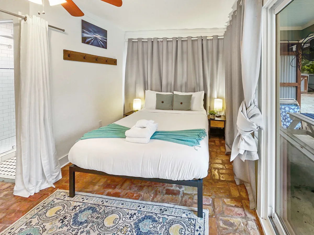 A neatly made bed with white and teal bedding in a room with white walls, tiled floor, and curtains.