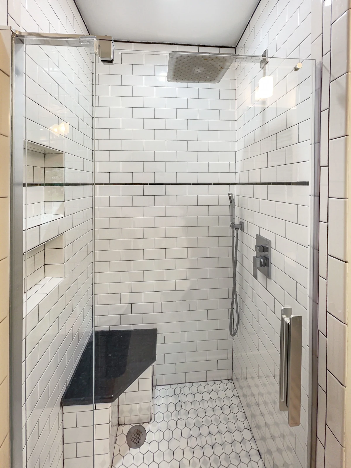 A clean and modern bathroom shower with white subway tiles, a black bench, and a rainfall showerhead.