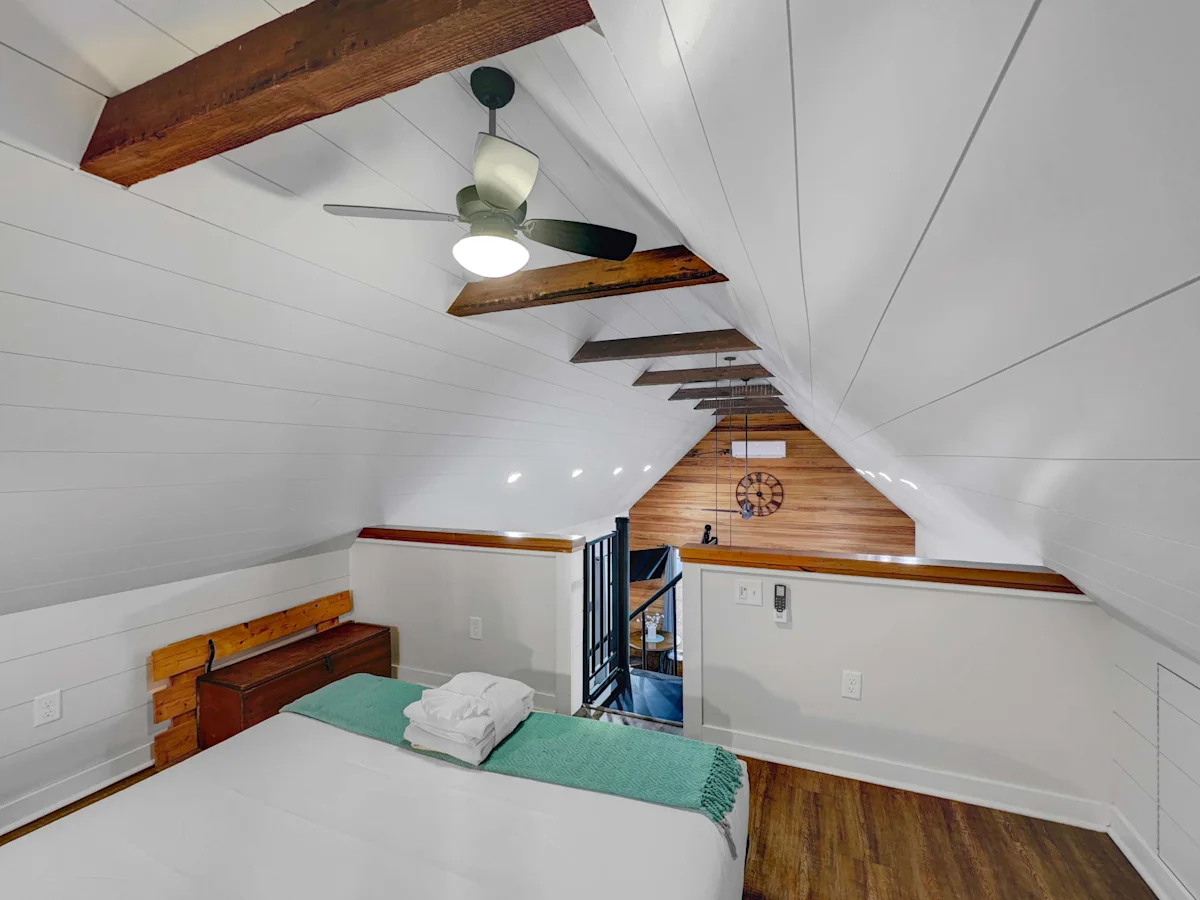 A cozy attic room with a twin bed, wooden beams, and a ceiling fan.
