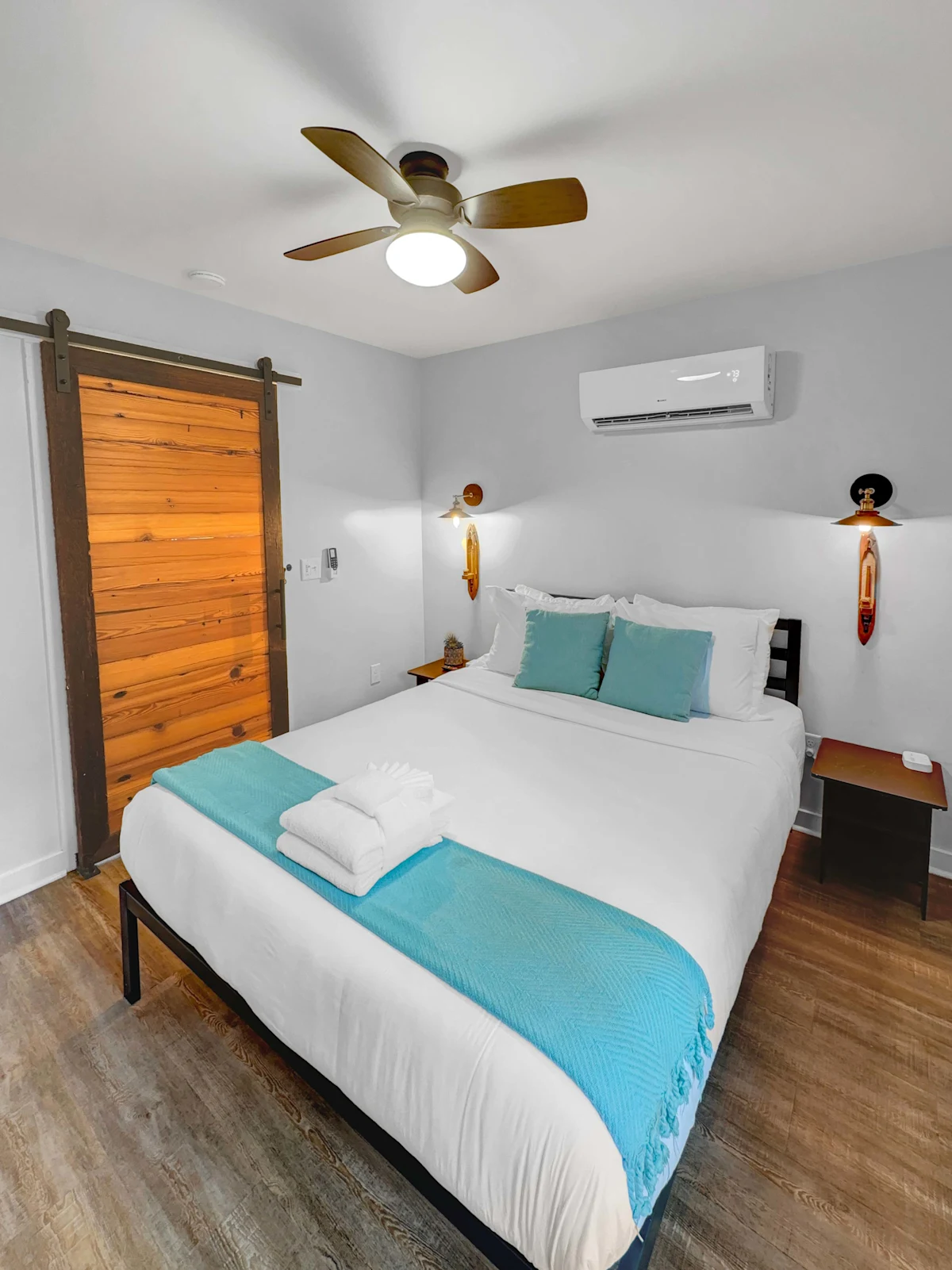 A neatly made hotel room with a large bed, wooden floor, sliding barn door, ceiling fan, and air conditioning unit.