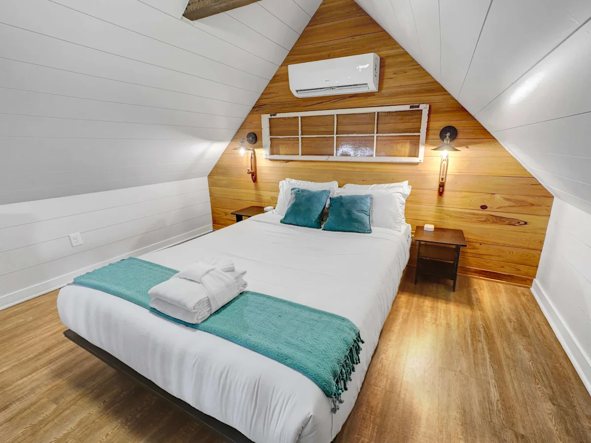 A neatly made bed with white sheets and teal accents in a cozy attic room with wood paneling and a vaulted ceiling.