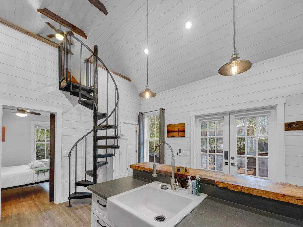 A small kitchen area with a sink, spiral staircase, and doorway to a bedroom