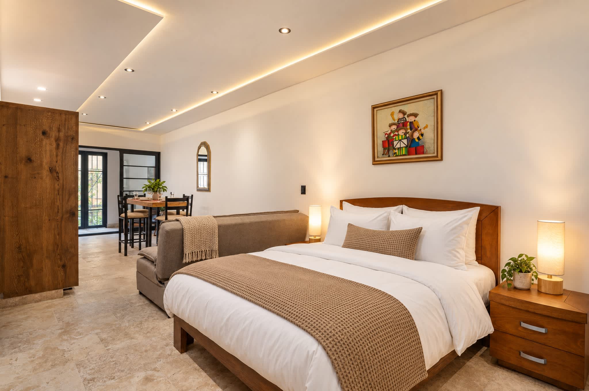 Recámara con cama matrimonial, cabecera de madera e iluminación cálida, que abre a la sala con comedor