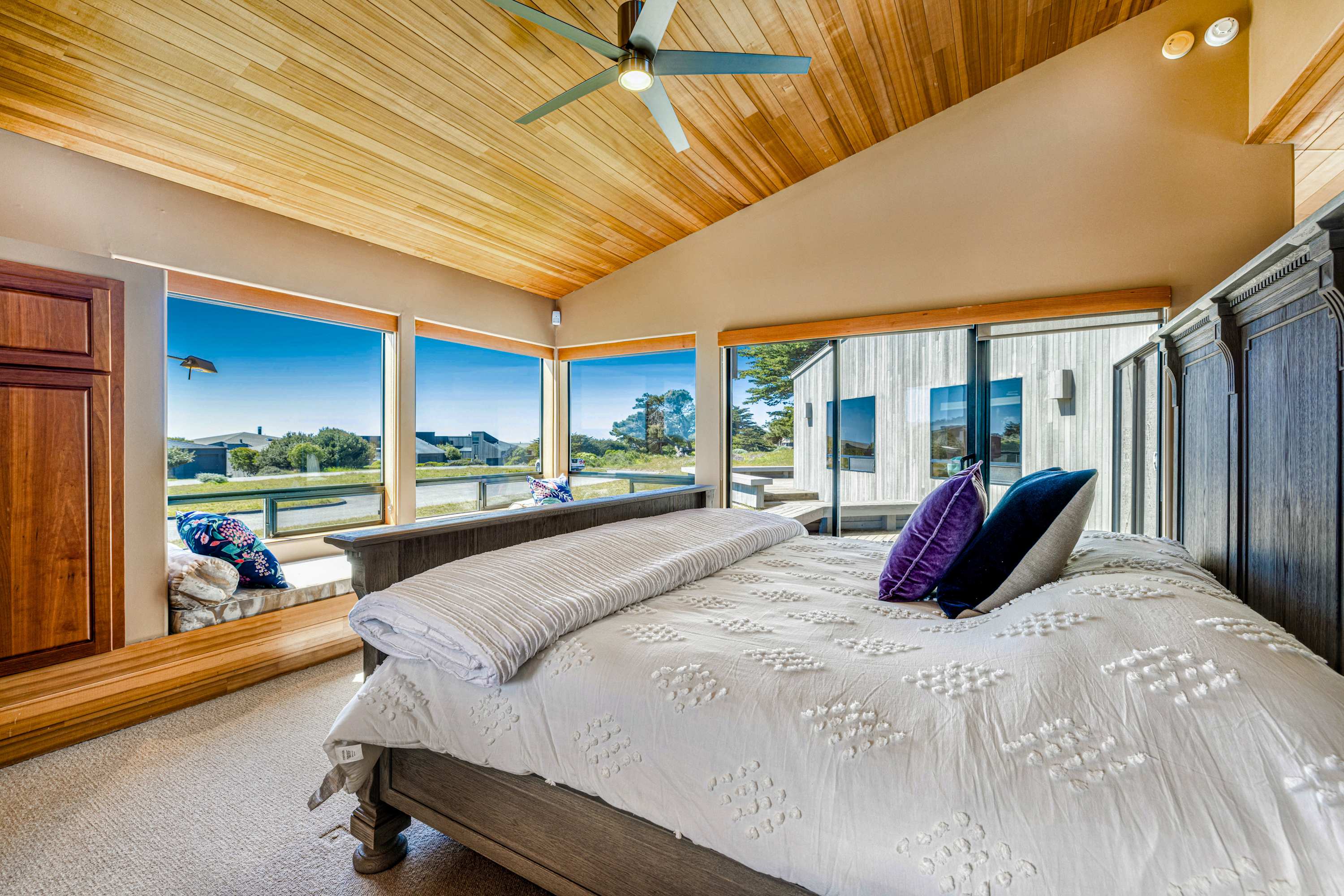 Purple velvet pillows against white tufted bedding — vaulted wood ceiling and a ceiling fan keep the primary room airy.