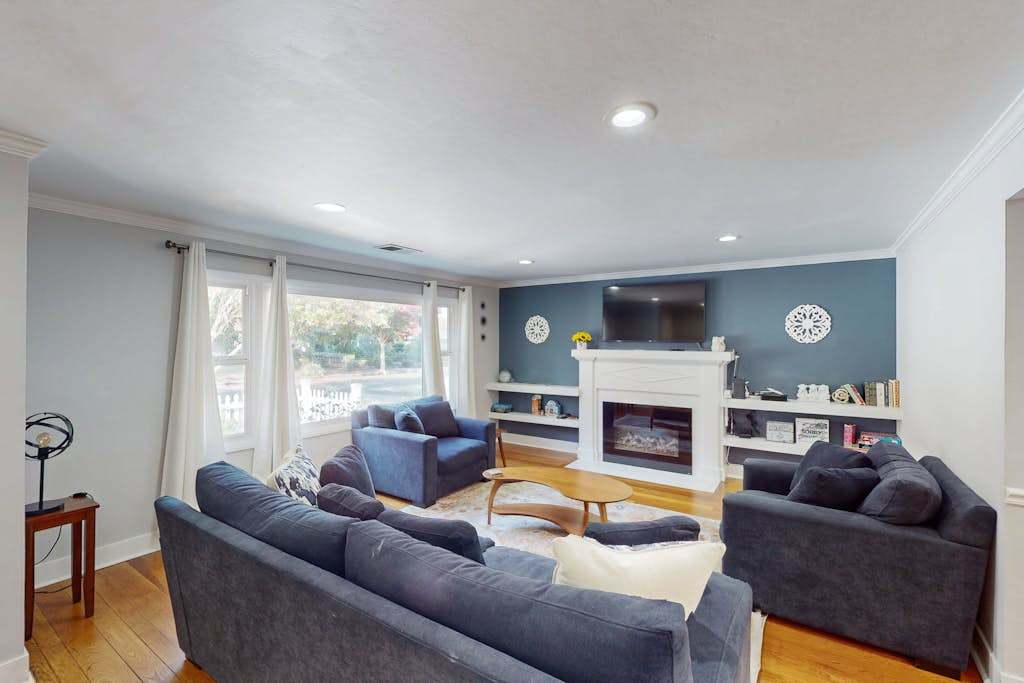 Gather around the fireplace in a sunlit living room with a bold blue accent wall, wall-mounted TV, built-in shelving, and plenty of plush seating for your group
