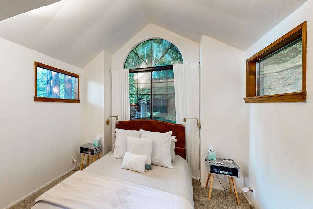 Rest easy in your serene bedroom — a queen bed with crisp white linens sits beneath a stunning arched window that frames the redwood canopy above Monte Rio.