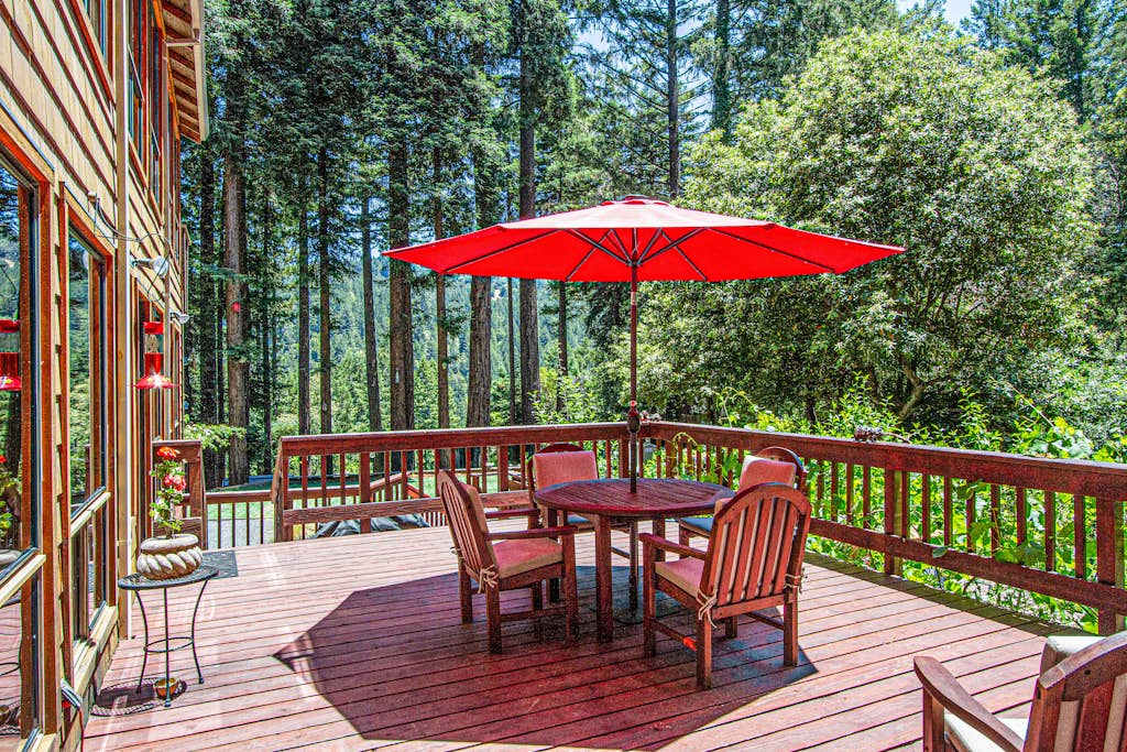 Savor your morning coffee on the spacious redwood deck — a shaded dining table for four awaits under a vibrant umbrella with forest canopy views all around.