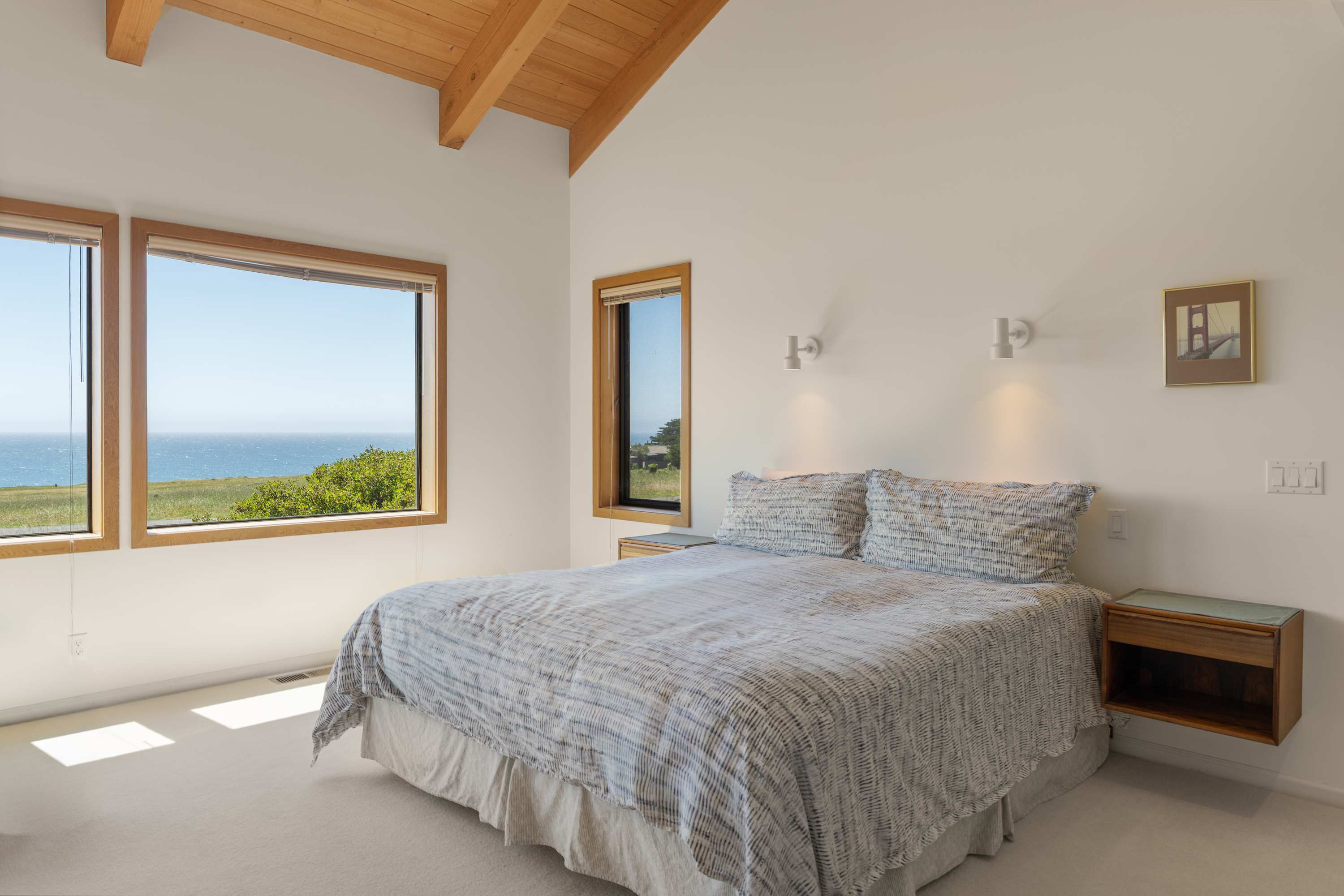 Wake on a king under the same vaulted beams — corner windows frame the meadow and ocean before you've even reached for coffee.