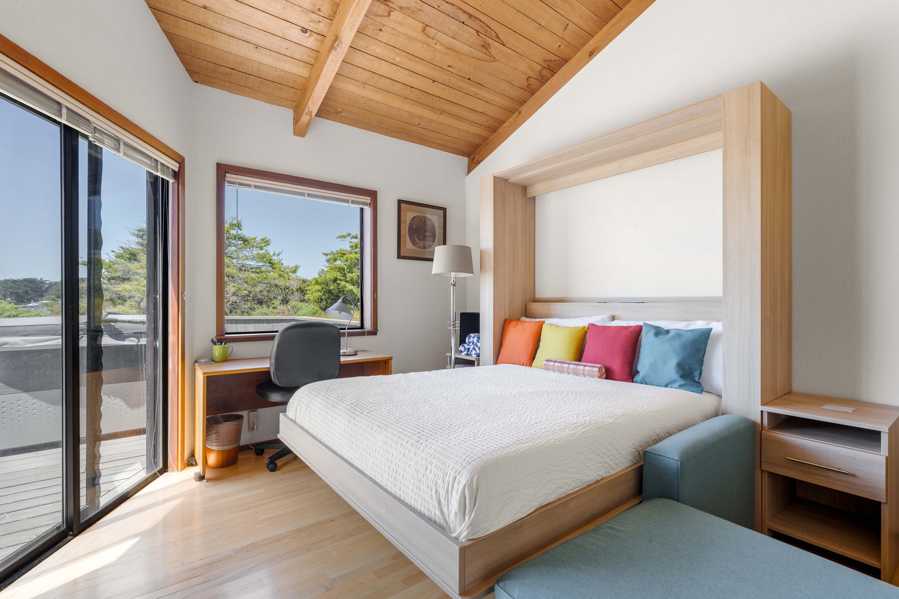 Bright bedroom with vaulted wood ceiling, king bed with colorful pillows, work desk, and sliding glass doors to a deck.
