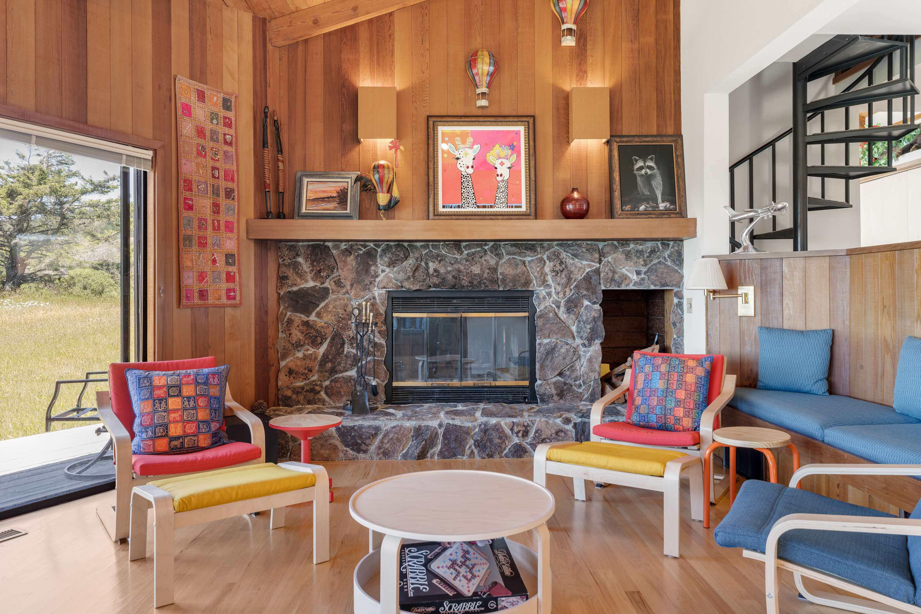 Cozy living room with a stone fireplace, eclectic art, and colorful seating overlooking a grassy meadow.