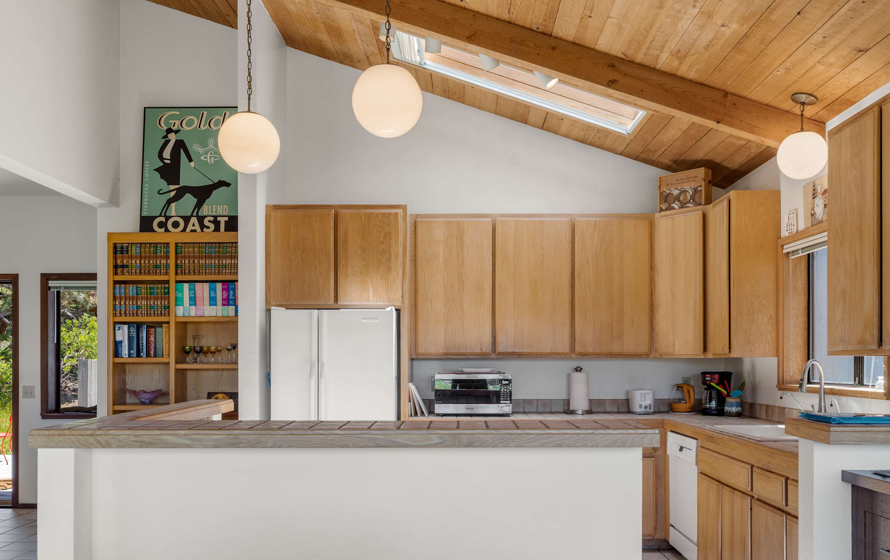 Bright kitchen with vaulted wood-beam ceiling, skylight, globe pendant lights, and oak cabinetry opening to a cozy bookshelf nook.