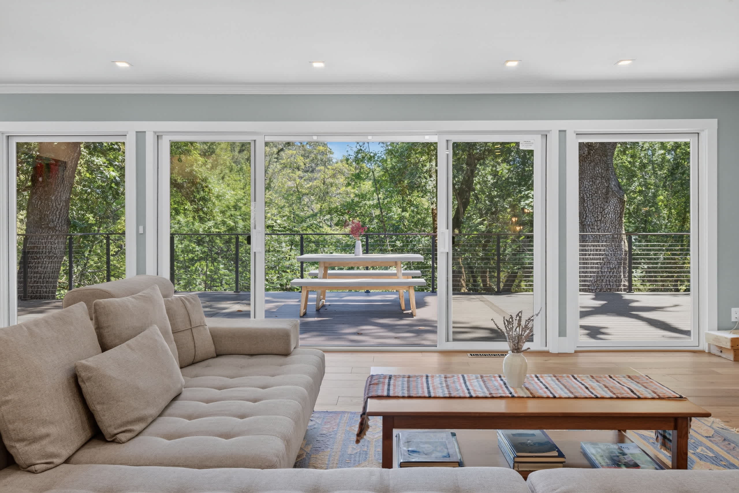 Sink into the linen sectional as oak canopies fill the wall of sliders — your deck and dining bench wait just beyond the glass.