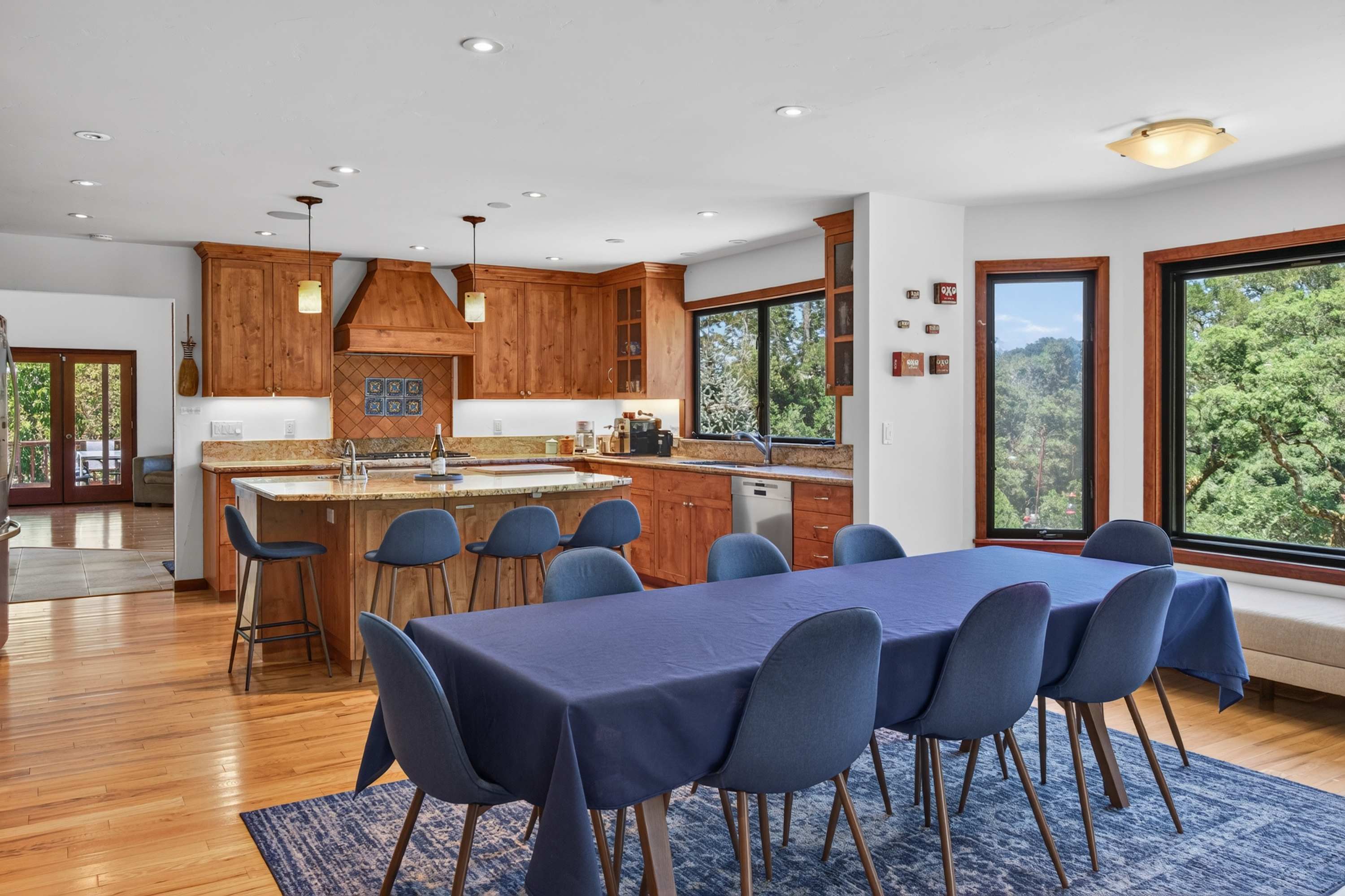 Pendants glow over the granite counters and the long table runs toward the window bench — 16 seats for a holiday dinner with the trees as backdrop.
