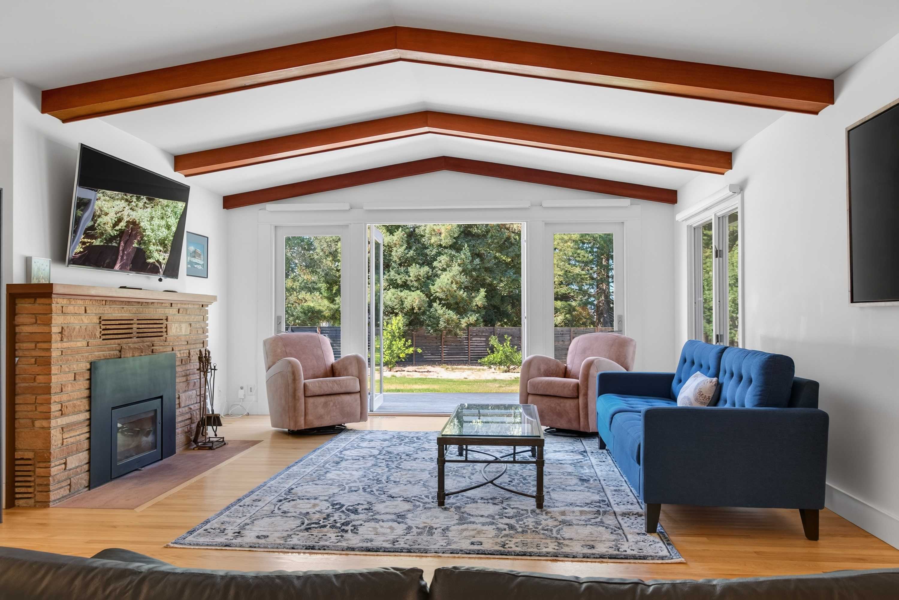 Vaulted beams and a brick fireplace anchor the great room, with glass doors framing the green yard beyond the blue sofa and patterned rug.