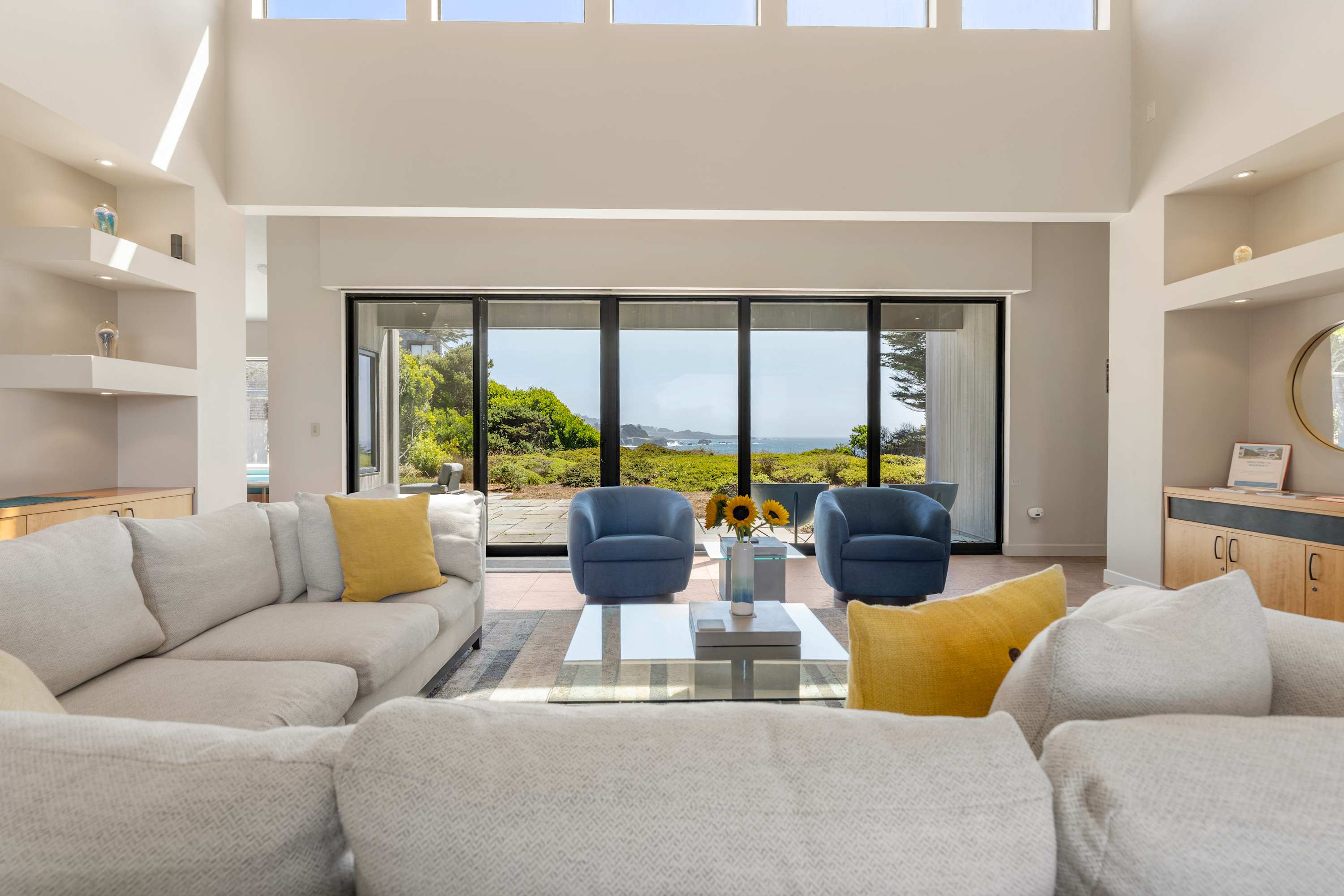 At Seaspray the living room is almost all glass — pale sectional, navy chairs, and the ocean sitting quietly past the meadow.