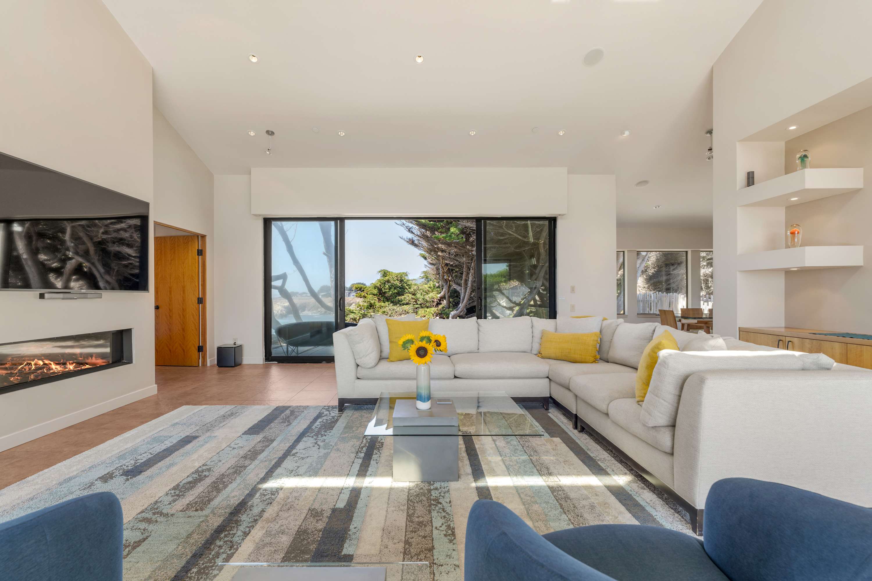 A long ribbon of fire under the TV, sunflower stems on the glass table, and those blue chairs pointed at the view.