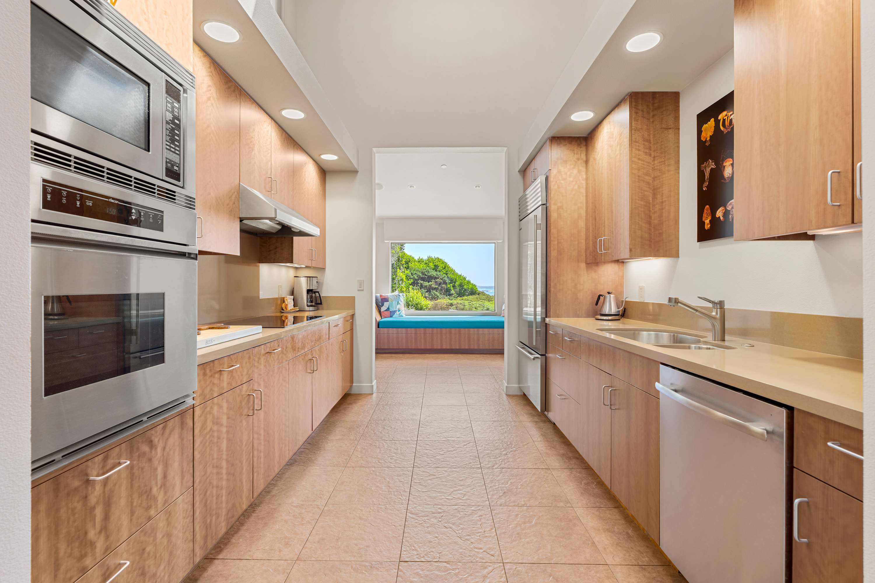 The galley runs toward a teal window seat. Cooktop, wall ovens, quartz — and a last scrap of ocean at the far end.