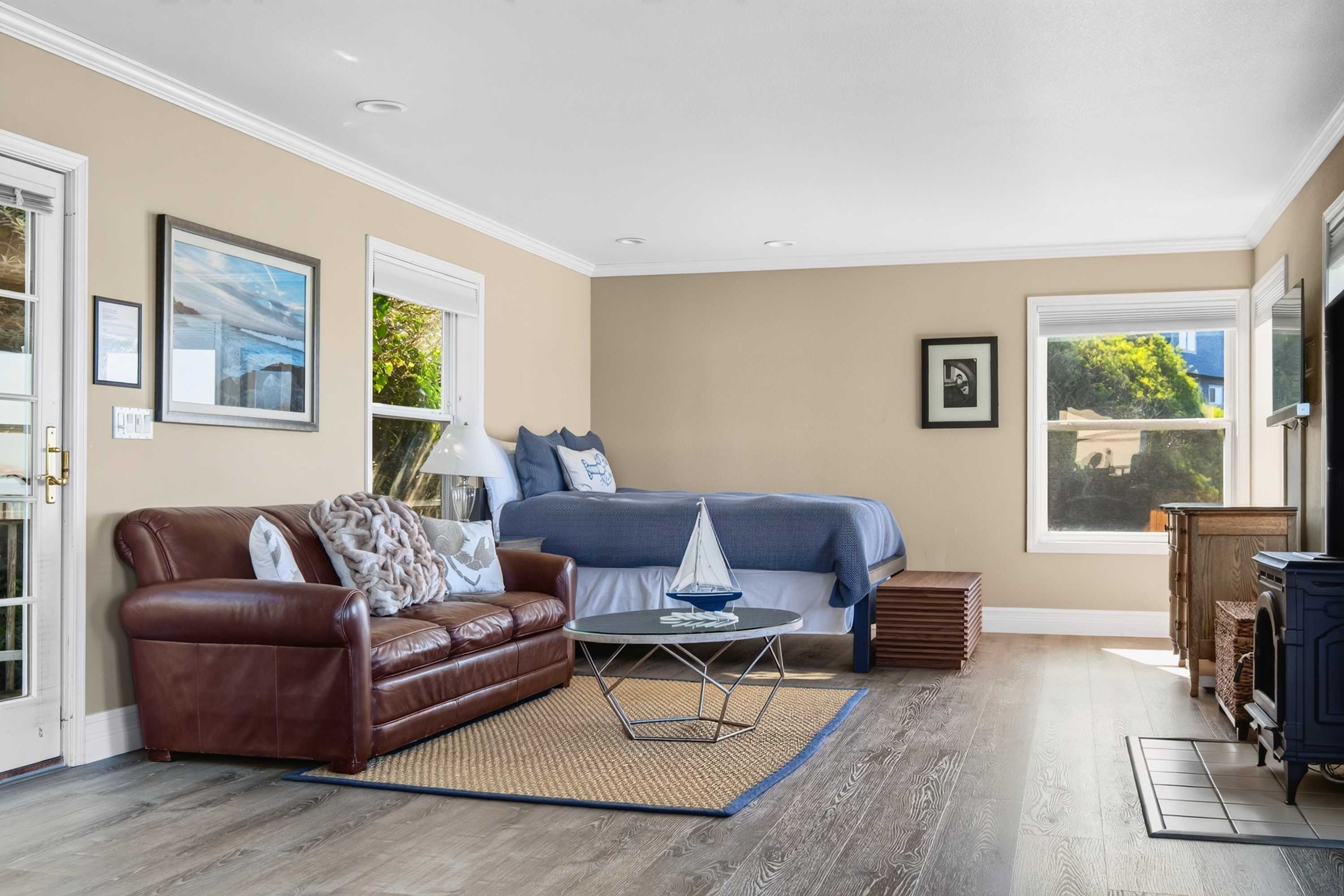 One open room holds the bed, brown leather sofa, wood stove, and jute rug — studio living steps from the water at Bayside Cottage.