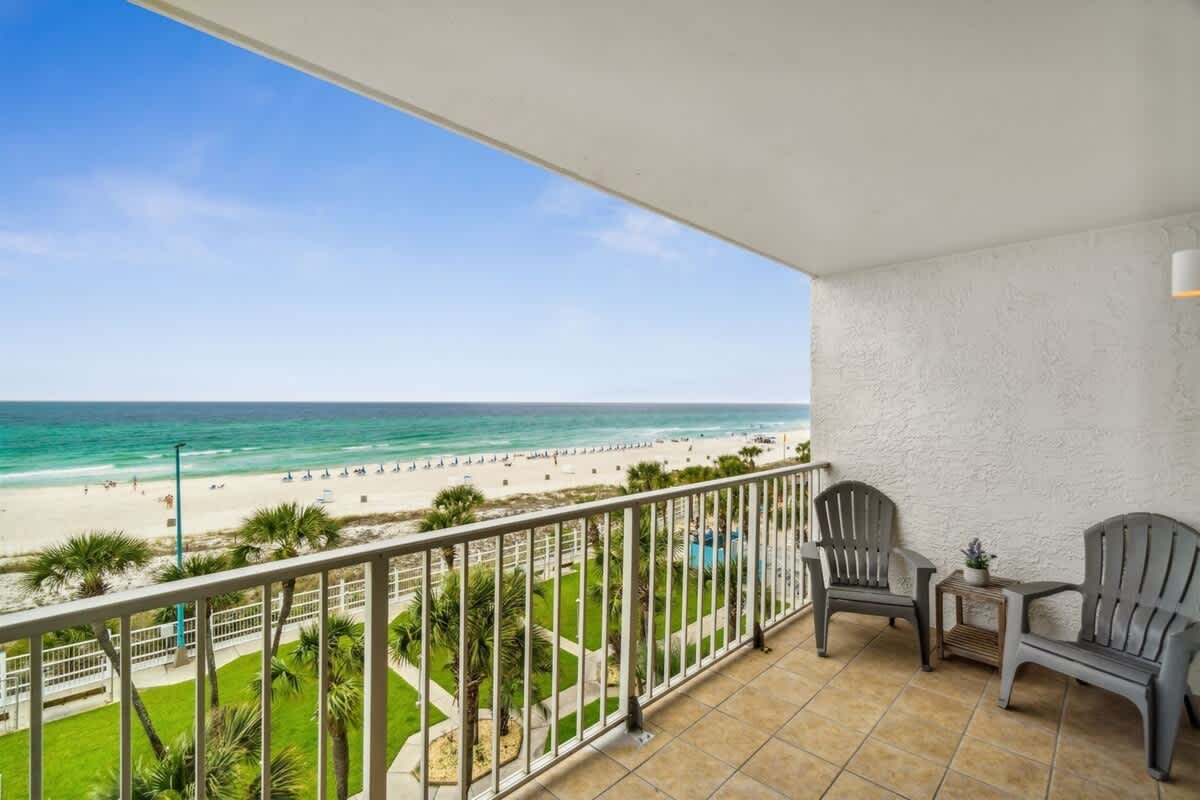 The Summit 531 – Balcony view of beach with two gray chairs, green grass, and palm trees.