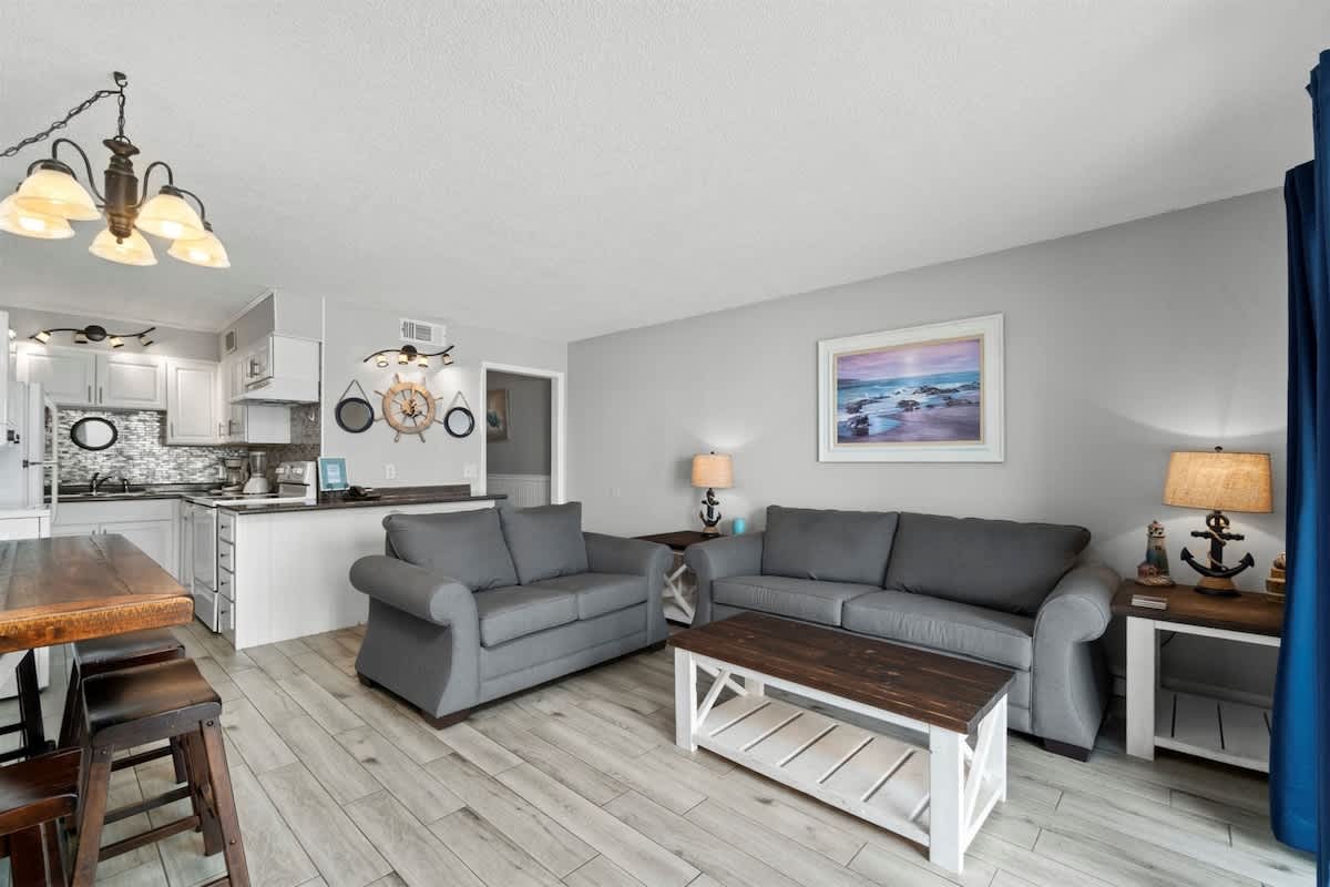 The Summit 728 – Living room with gray sofas, wooden coffee table, dining area, and kitchenette. Tropical-themed decor and window.