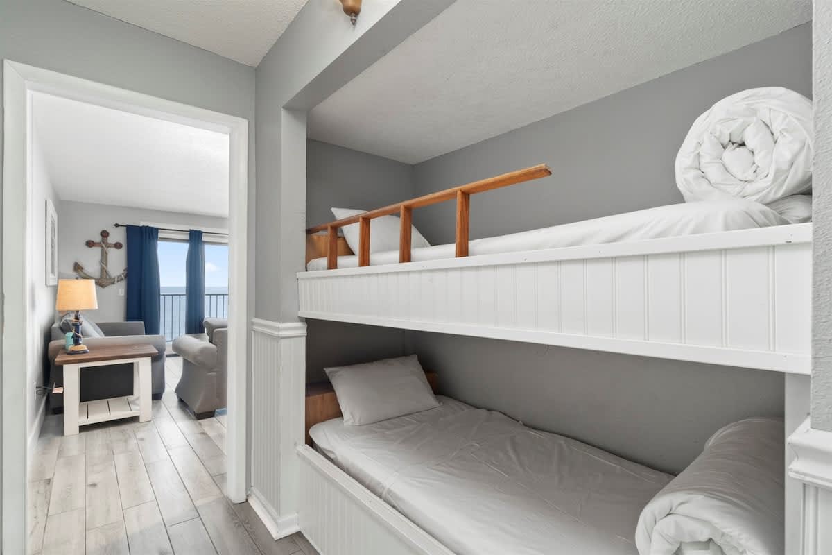 The Summit 728 – Bunk beds with white bedding and a living area view through a doorway.